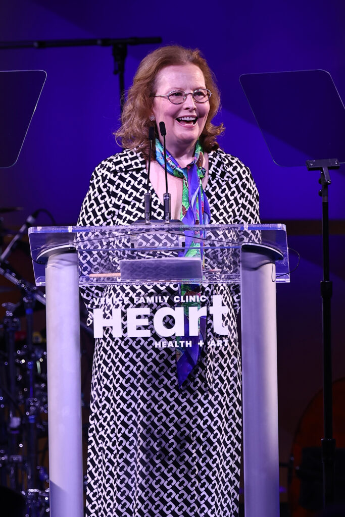A Woman Wearing Glasses And A Patterned Dress Stands At A Clear Podium With Heart Written On It, Smiling While Speaking Into A Microphone At An Event With Purple Lighting.