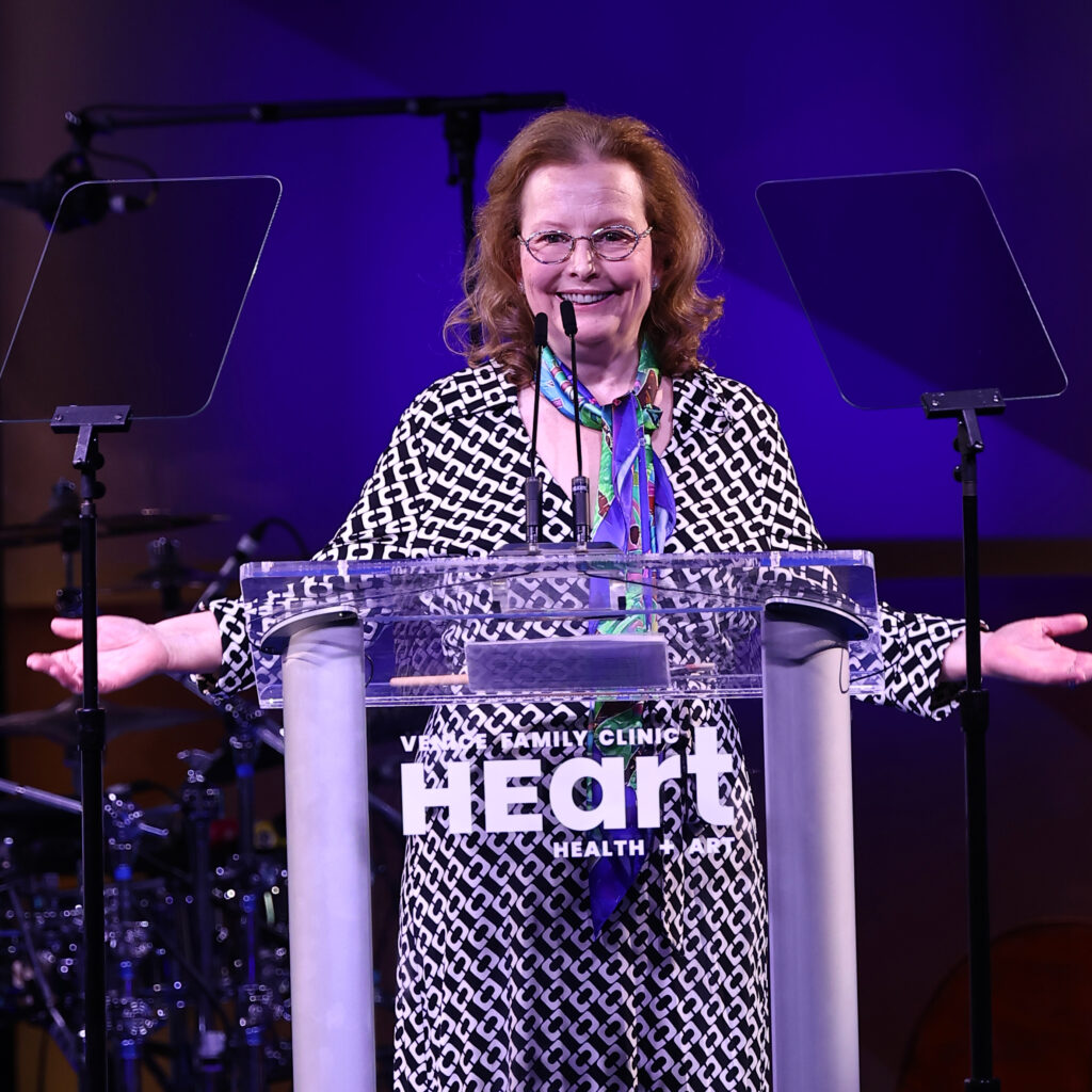 A Smiling Woman With Glasses Stands At A Clear Podium With Heart Health Written On The Front, Arms Outstretched, On A Stage With Purple Lighting. Two Transparent Screens Are Positioned On Either Side Of Her.