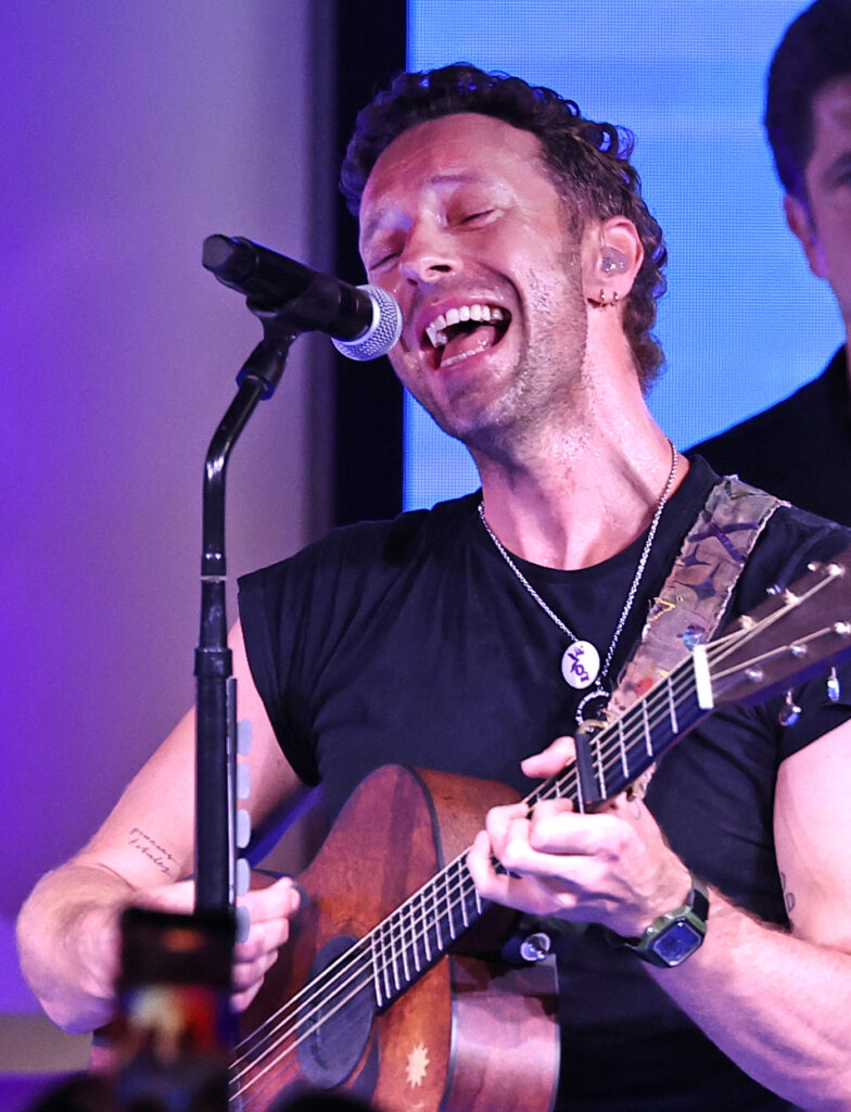 A Man Passionately Sings And Plays An Acoustic Guitar On Stage, Wearing A Black Shirt And Necklace, With Stage Lights Illuminating Him From The Side.