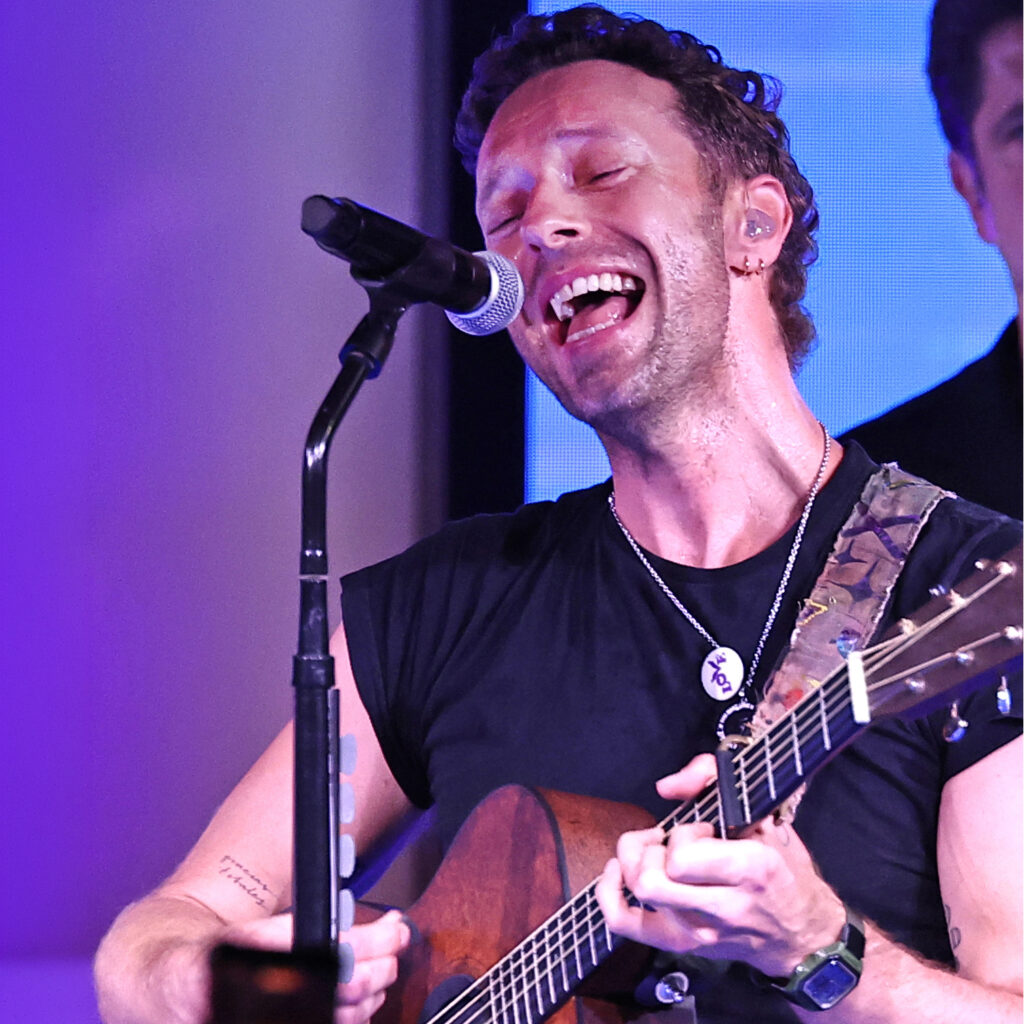 A Musician Passionately Sings Into A Microphone While Playing An Acoustic Guitar On Stage, Wearing A Black T-shirt And Necklace, With Purple And Blue Stage Lighting In The Background.