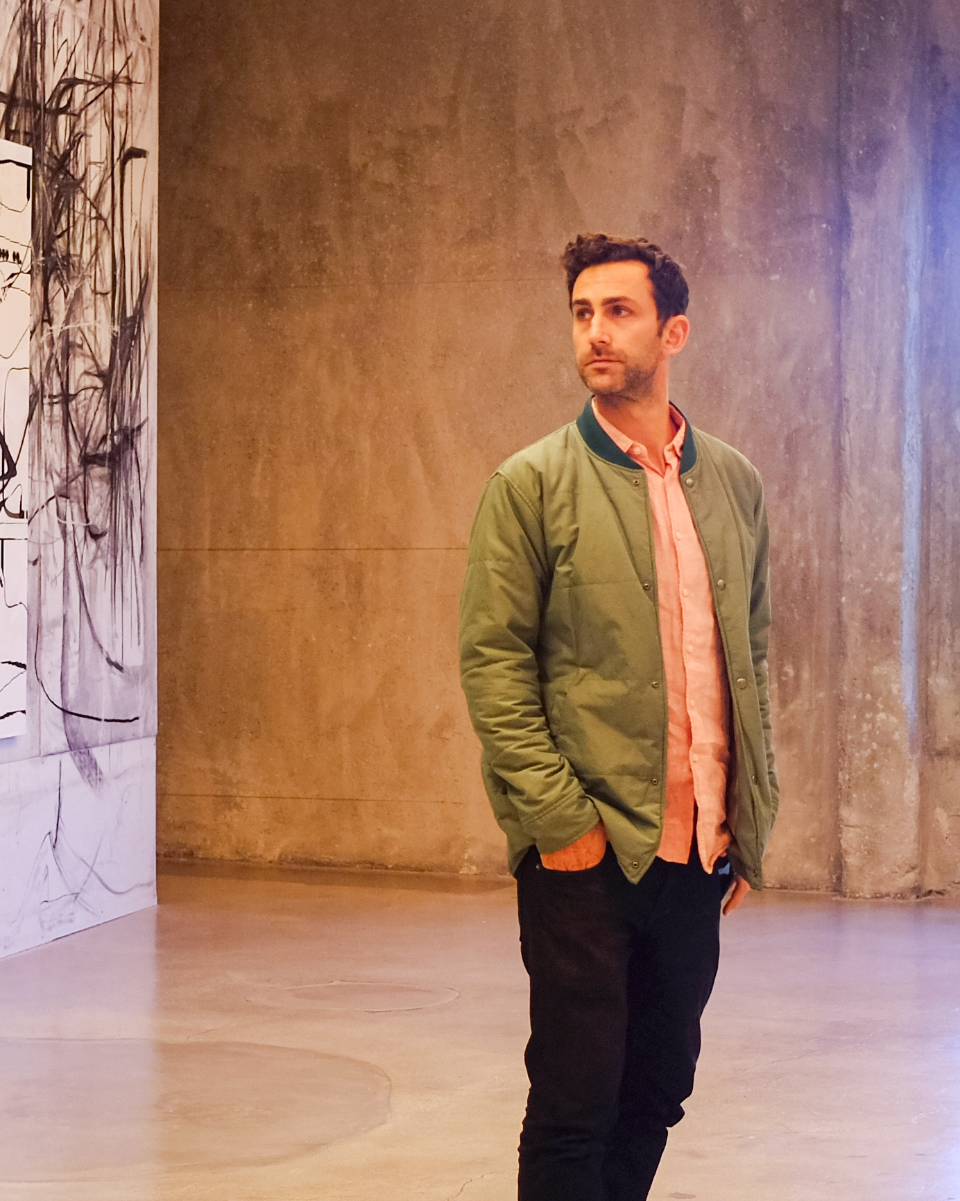 A Man Wearing A Green Jacket, Pink Shirt, And Black Pants Stands Indoors On A Polished Concrete Floor Near A Wall With Abstract Black-and-white Artwork, Looking Thoughtfully To The Side. A Man Wearing A Green Jacket, Pink Shirt, And Black Pants Stands Indoors On A Polished Concrete Floor Near A Wall With Abstract Black-and-white Artwork, Looking Thoughtfully To The Side.