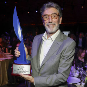A Man With Gray Hair, A Beard, And Glasses Smiles While Holding A Blue And Silver Trophy That Reads “Chuck Lorre, Lifetime Achievement Award, HE.ART Gala 2024.” People Are Seated At Tables In The Background.
