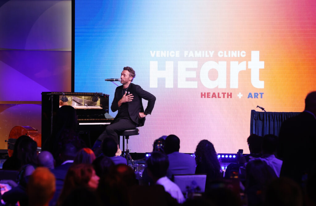 A Musician Sits At A Piano On Stage, Performing For An Audience At The Venice Family Clinic Heart Health + Art Event. A Large Screen Behind Him Displays The Events Name In Bold Letters.