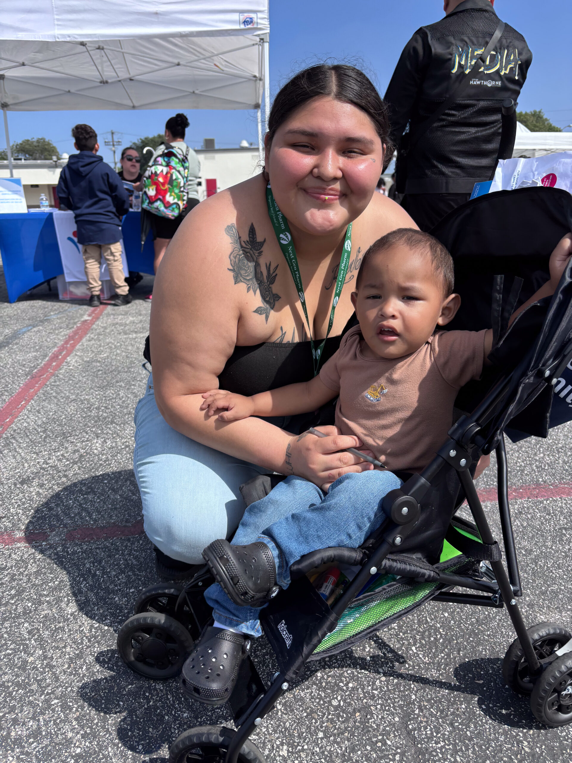 A Woman With Long Dark Hair Kneels Next To A Toddler Sitting In A Stroller At An Outdoor Hawthorne Event. She Smiles At The Camera, Holding The Child, While People And Booths Are Visible In The Background. A Woman With Long Dark Hair Kneels Next To A Toddler Sitting In A Stroller At An Outdoor Hawthorne Event. She Smiles At The Camera, Holding The Child, While People And Booths Are Visible In The Background.