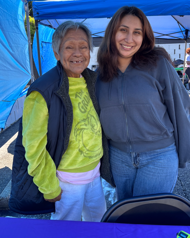 Two Women Stand Side By Side Under A Blue Canopy, Smiling At The Camera. One Wears A Green Grinch Sweatshirt And Black Vest, While The Other Wears A Gray Zip-up Hoodie And Jeans. A Purple Table Is In The Foreground.
