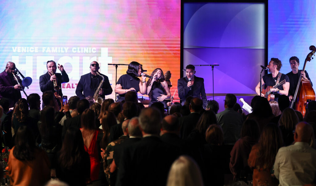 A Band Performs Live On Stage With Multiple Singers And Musicians In Front Of A Large Seated Audience At An Indoor Event With Colorful Lights And A Venice Family Clinic Sign In The Background.