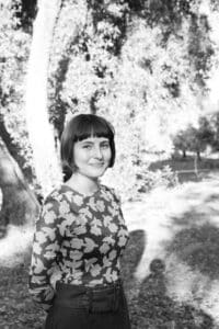 A Young Woman With A Bob Haircut And Patterned Shirt Stands Outdoors, Smiling Slightly. Sunlight Filters Through The Trees, Casting Shadows On The Ground In A Natural, Park-like Setting. The Image Is In Black And White.