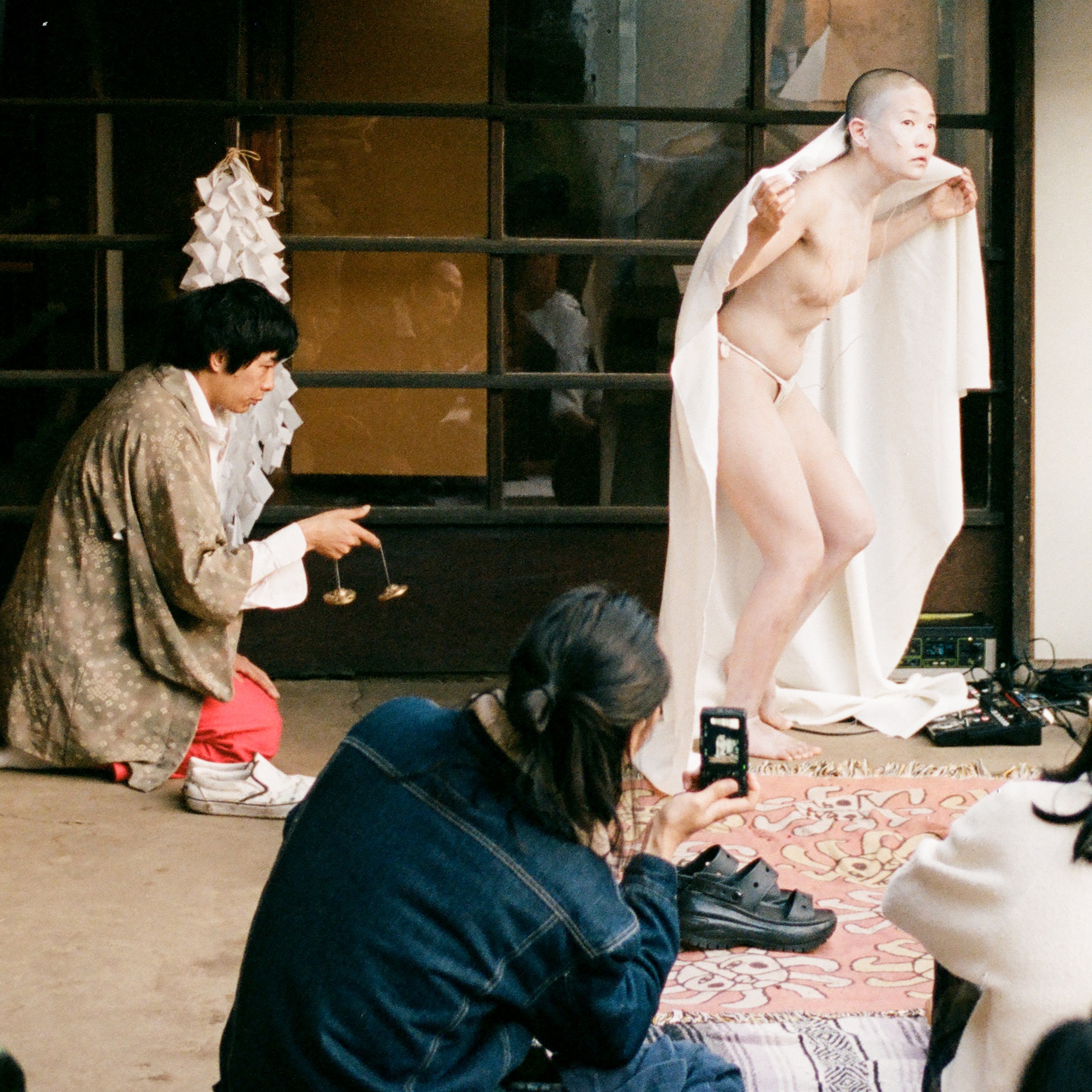A Performer Draped In A White Sheet Stands Barefoot, Posing Next To A Person Kneeling And Holding Cymbals. Several People Sit Watching, One Capturing The Scene With A Phone. The Background Features Large Windows.
