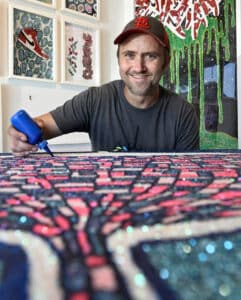 A Smiling Man In A Red-and-black Cap Applies Blue Paint To A Colorful, Mosaic-style Artwork. Hes In A Gallery Space With Framed Art On White Walls And A Large, Drip-style Painting Behind Him.