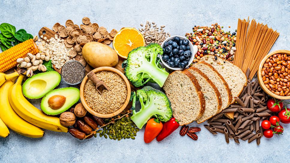 A Colorful Assortment Of Healthy Whole Foods, Including Fruits, Vegetables, Grains, Seeds, Nuts, Pasta, Bread, And Legumes, Arranged On A Light Blue Surface.