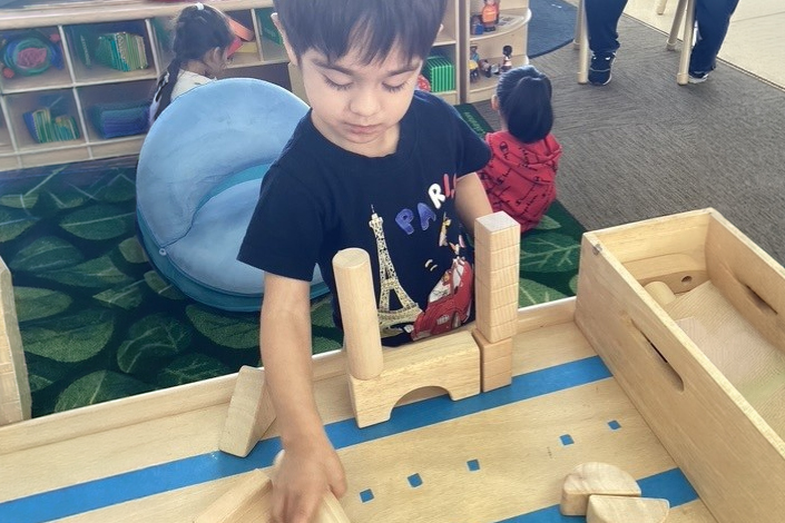 A Young Child Wearing A Black T-shirt Builds A Structure With Wooden Blocks On A Table In A Classroom, With Shelves, Toys, And Other Children Visible In The Background.