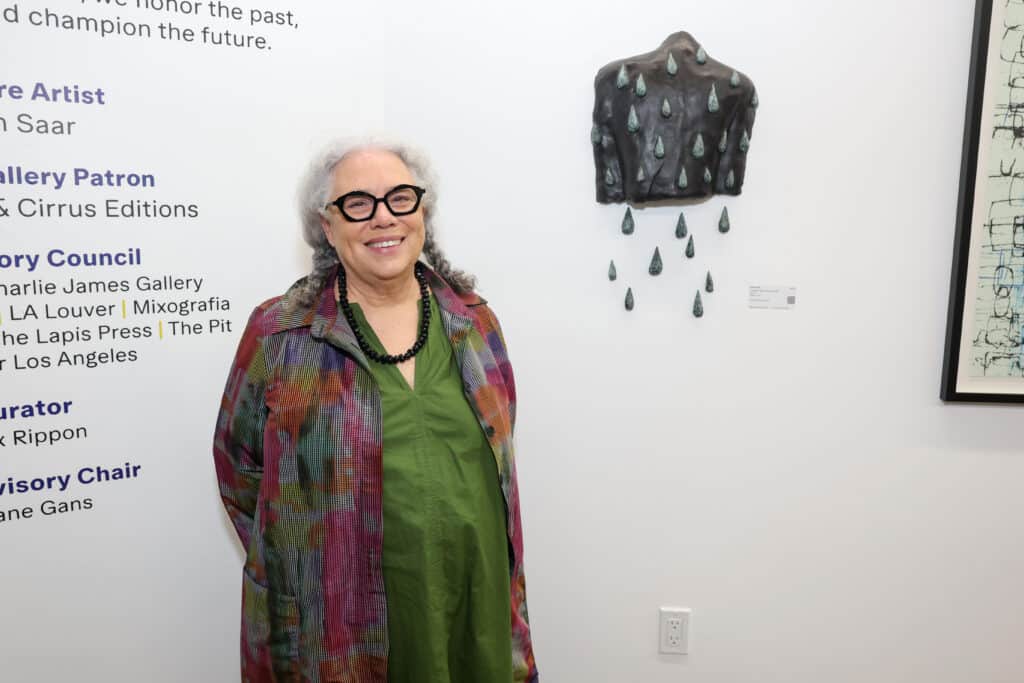 An Older Woman With Gray Hair, Glasses, And A Colorful Coat Over A Green Dress Stands Smiling In An Art Gallery Next To A Mixed-media Artwork Resembling A Dark Shirt With Raindrops Attached To A White Wall.