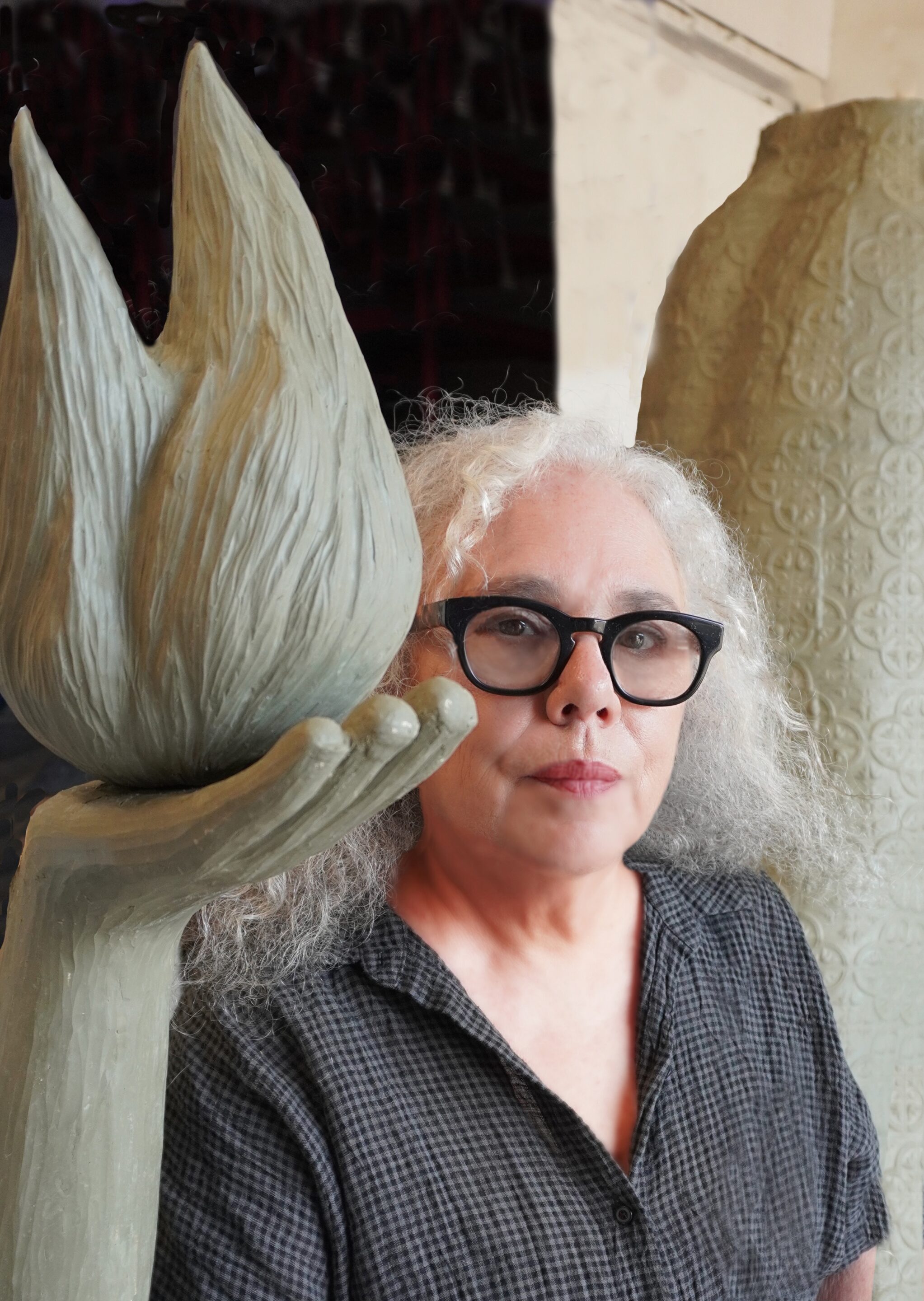 A Person With Curly Gray Hair And Black Glasses Stands Beside A Large, Abstract Sculpture Of A Hand Holding A Flame-shaped Object And Another Textured Column. They Wear A Dark, Checkered Shirt And Have A Serious Expression.