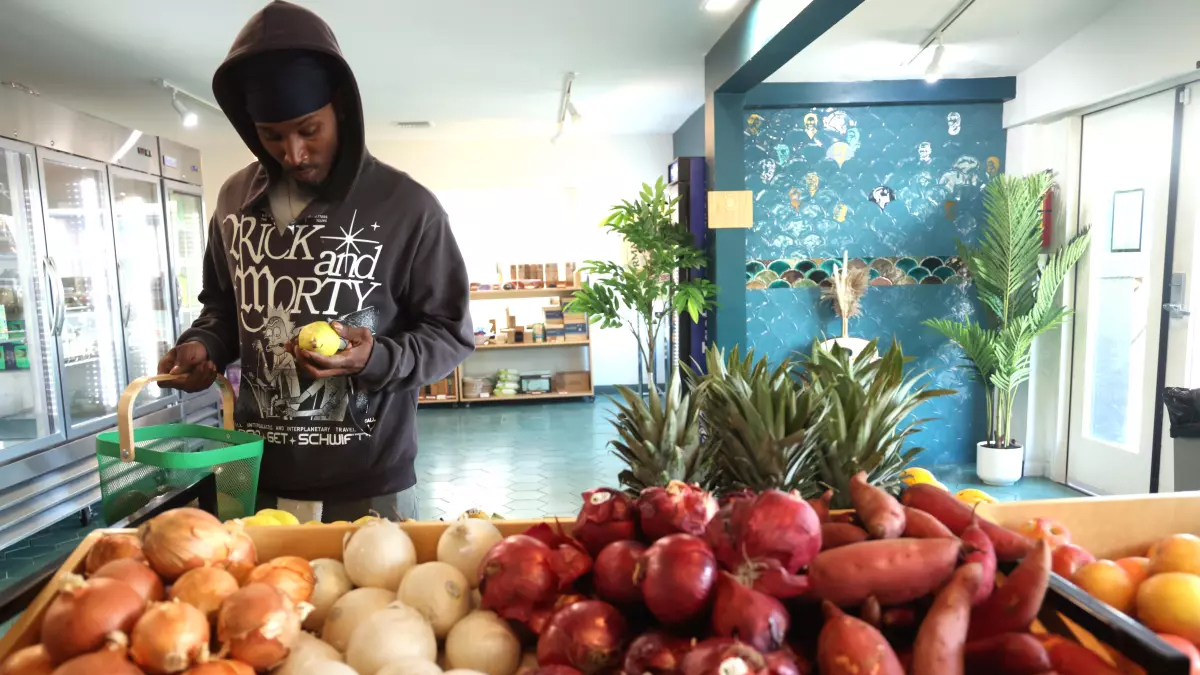 A Person In A Hoodie Examines An Onion In A Grocery Store, Standing Beside A Basket, With A Display Of Fresh Produce In The Foreground And A Colorful, Plant-filled Interior In The Background. A Person In A Hoodie Examines An Onion In A Grocery Store, Standing Beside A Basket, With A Display Of Fresh Produce In The Foreground And A Colorful, Plant-filled Interior In The Background.