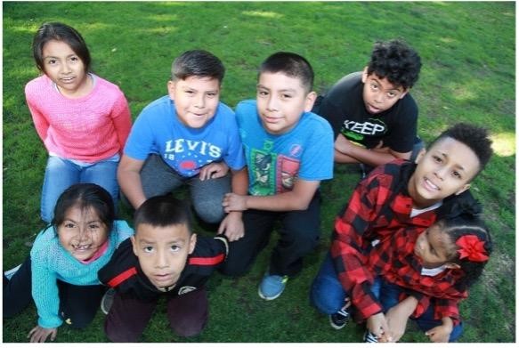 Eight Children Sit And Kneel On Grass, Looking Up And Smiling At The Camera. They Wear Colorful Casual Clothing And Appear To Be Enjoying Time Together Outdoors.