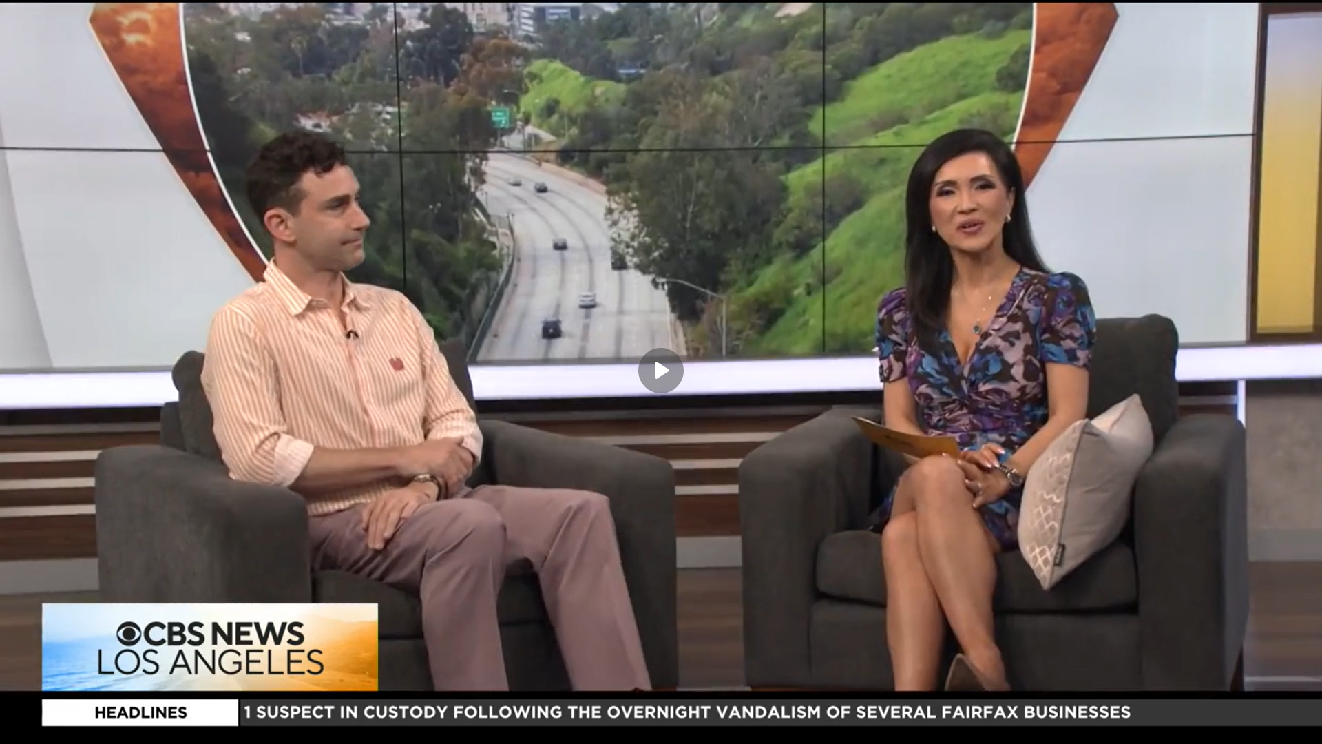 A man and a woman sit on armchairs during a CBS News Los Angeles segment. Behind them is a screen displaying a green, hilly landscape with a road. The woman holds a notepad, and both are engaged in conversation.
