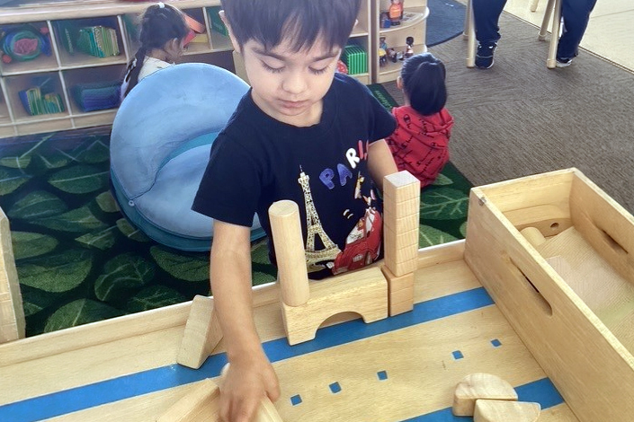 A Young Child Builds With Wooden Blocks At A Play Table In A Classroom. Other Children Are In The Background, And Shelves With Toys And Books Line The Wall. The Child Is Focused And Engaged In The Activity.