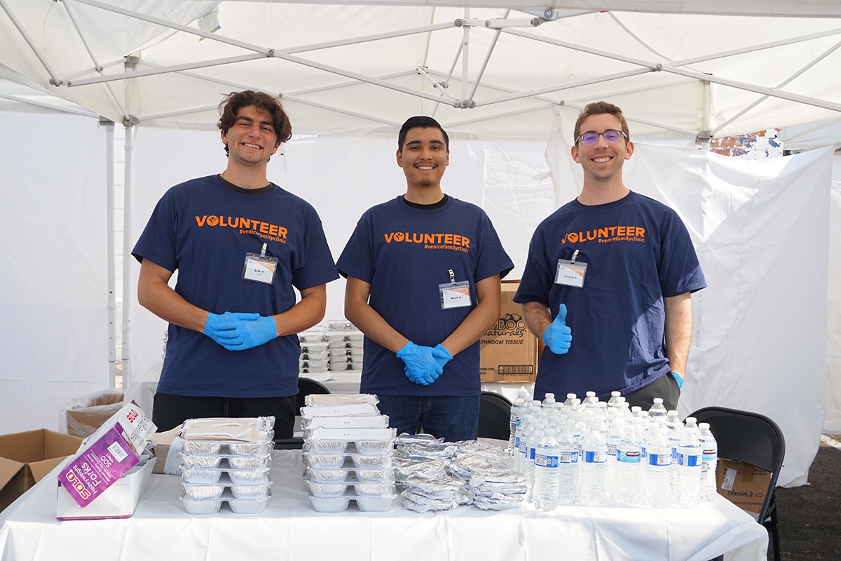 Three Volunteers Wearing Navy Blue VOLUNTEER Shirts And Gloves Stand Behind A Table With Stacked Meal Containers And Bottled Water Under A White Canopy Tent, Smiling At The Camera.