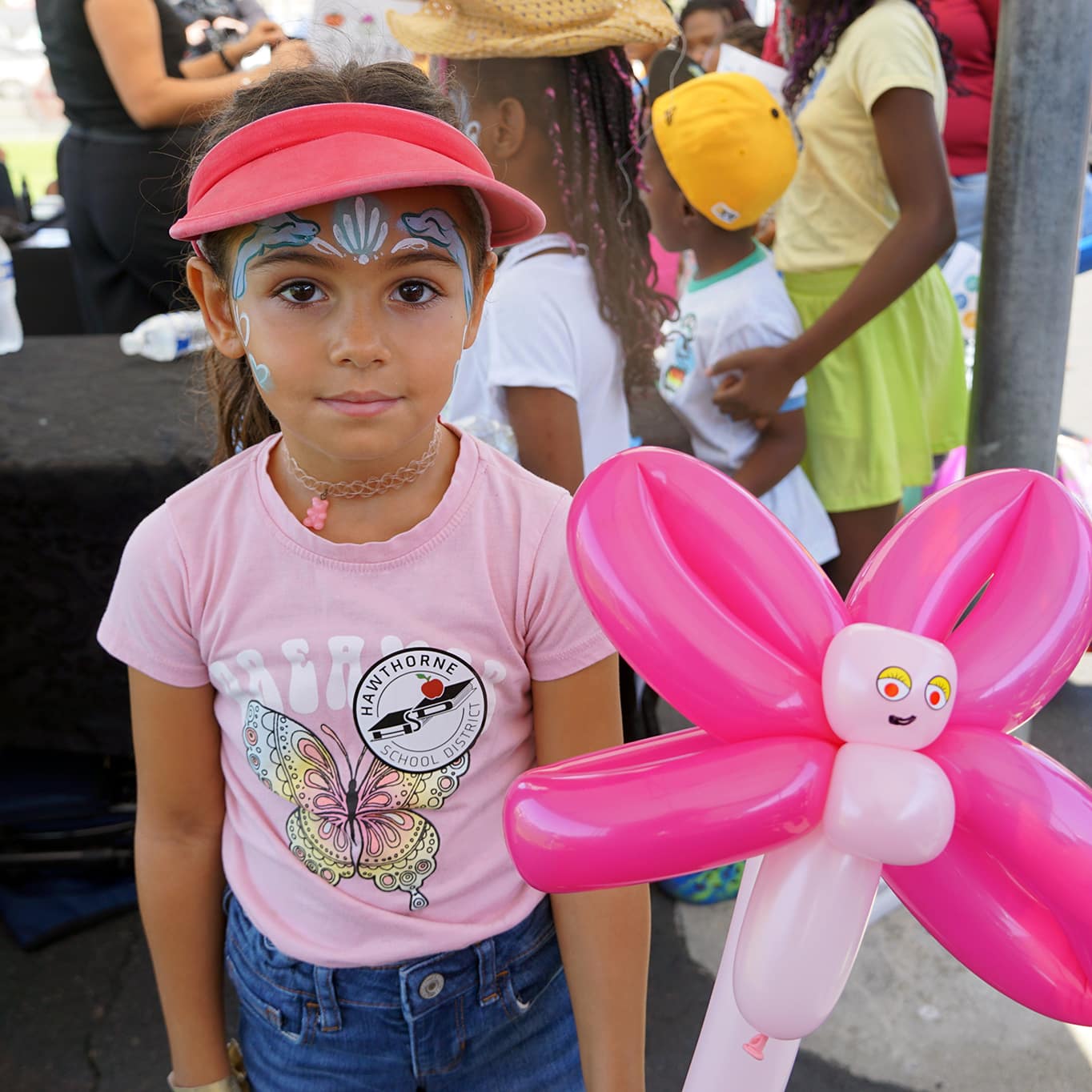 A Young Girl With Face Paint And A Pink Visor Holds A Pink Balloon Flower. She Wears A Pink T-shirt With A Butterfly Design, Standing Outdoors Among Other Children At An Event.