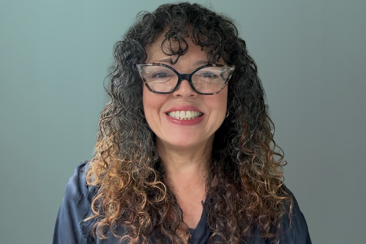 A Woman With Curly Brown Hair And Glasses Smiles At The Camera, Wearing A Dark Blouse And Standing In Front Of A Plain, Light-colored Background.