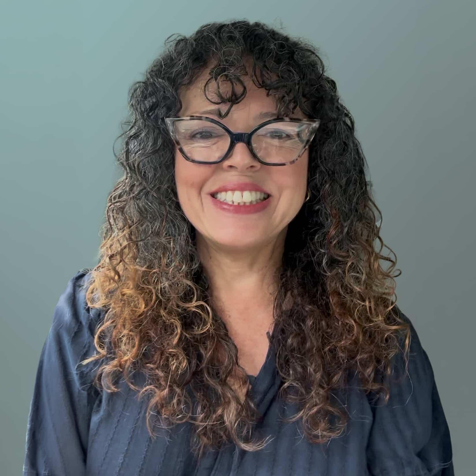 A Woman With Curly, Shoulder-length Hair And Glasses Is Smiling At The Camera. She Is Wearing A Dark Blouse And Standing In Front Of A Plain, Light Gray Background. A Woman With Curly, Shoulder-length Hair And Glasses Is Smiling At The Camera. She Is Wearing A Dark Blouse And Standing In Front Of A Plain, Light Gray Background.