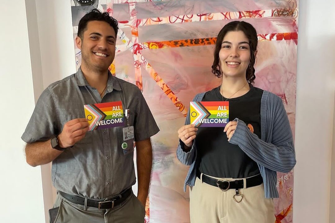 Two People Smiling And Holding Colorful Cards That Read All Are Welcome. They Stand In Front Of A Vibrant, Abstract Background With Various Colors And Patterns.