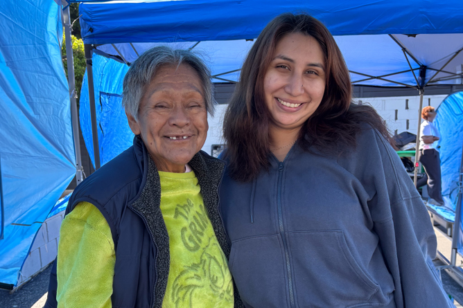 Two People Stand Smiling Under Blue Canopy Tents Outdoors. The Older Person On The Left Has Short Gray Hair And Wears A Yellow Shirt And Dark Vest, While The Younger Person On The Right Has Long Brown Hair And Wears A Gray Hoodie.