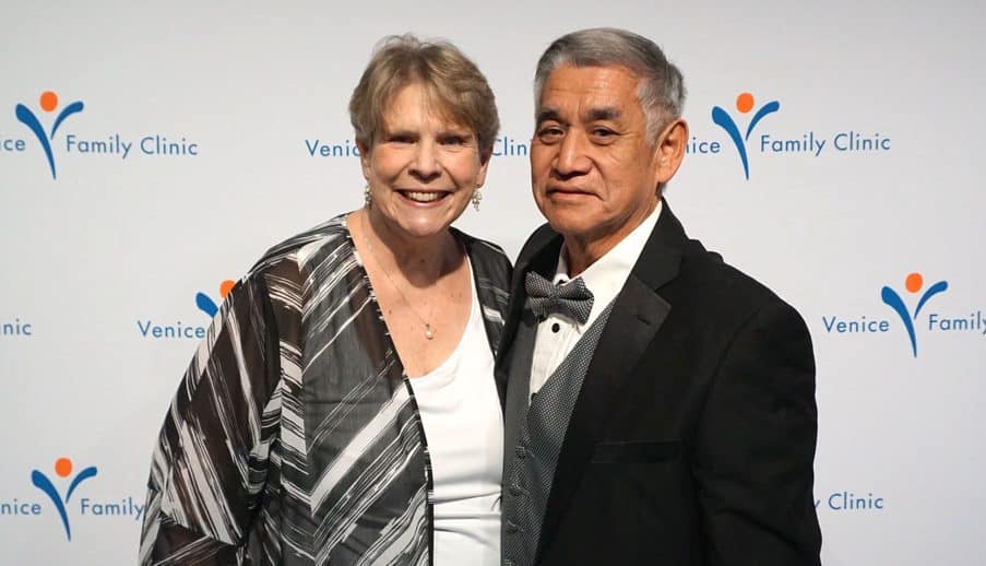 A Smiling Woman And Man Dressed In Formal Attire Pose Together In Front Of A Backdrop With The Venice Family Clinic Logo.