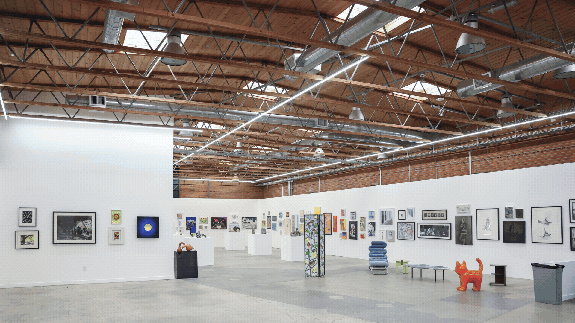 A Spacious Modern Art Gallery With High Wooden Ceilings, White Walls Displaying Various Framed Artworks, Sculptures On Pedestals, And Several Colorful Art Pieces And Furniture Scattered Around The Room. A Spacious Modern Art Gallery With High Wooden Ceilings, White Walls Displaying Various Framed Artworks, Sculptures On Pedestals, And Several Colorful Art Pieces And Furniture Scattered Around The Room.
