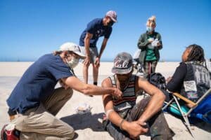 A Masked Healthcare Worker Examines A Seated Man On A Sunny Beach, While Others, Some Wearing Masks, Stand Or Sit Nearby. The Sky Is Clear And Blue.