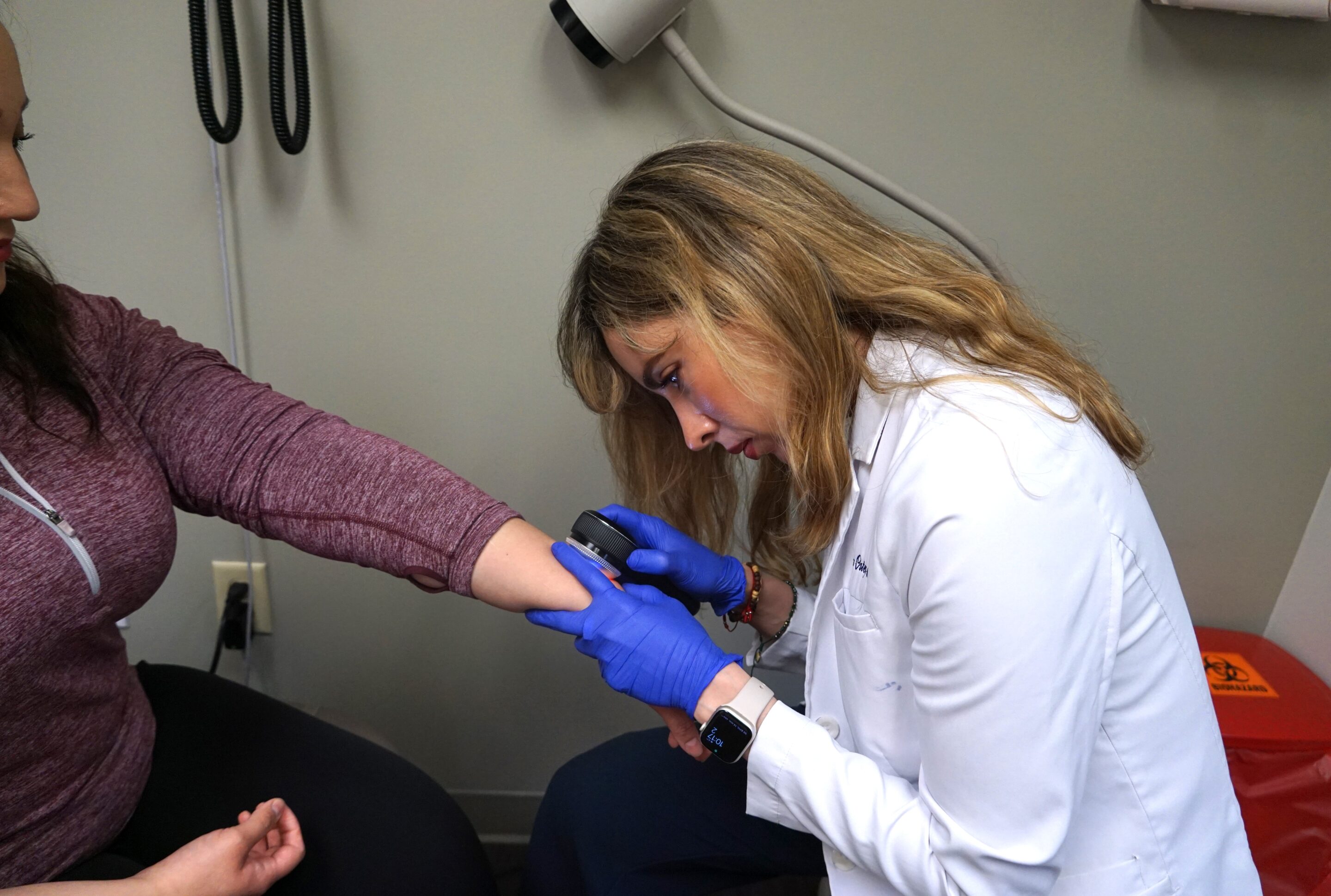 A Healthcare Professional Wearing Blue Gloves Examines A Patient’s Forearm With A Dermatoscope In A Medical Office. The Patient Is Seated And Has Their Sleeve Rolled Up For The Skin Check.