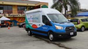 A Blue And White Venice Family Clinic Mobile Van Is Parked Outside St. Margaret’s Center, Near Other Vehicles And A Palm Tree. The Clinic Offers Medical Services To The Local Community.