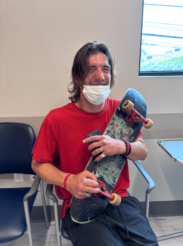Smiling Person With Long Hair Wearing A Red Shirt And A Partially Lowered surgical Mask Sits In A Chair Holding A Skateboard. There Is A Window, Another Chair, And A Small Table In The Room.