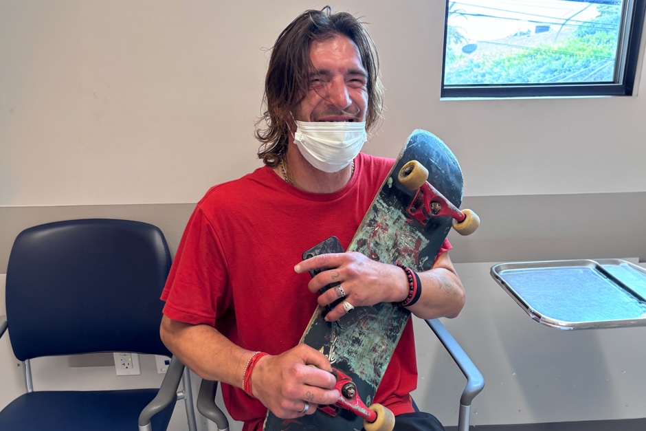 A Smiling Man In A Red Shirt Sits Indoors, Wearing A Face Mask And Holding A Skateboard On His Lap. He Is Seated Next To Empty Chairs And A Window, With Sunlight Coming In.