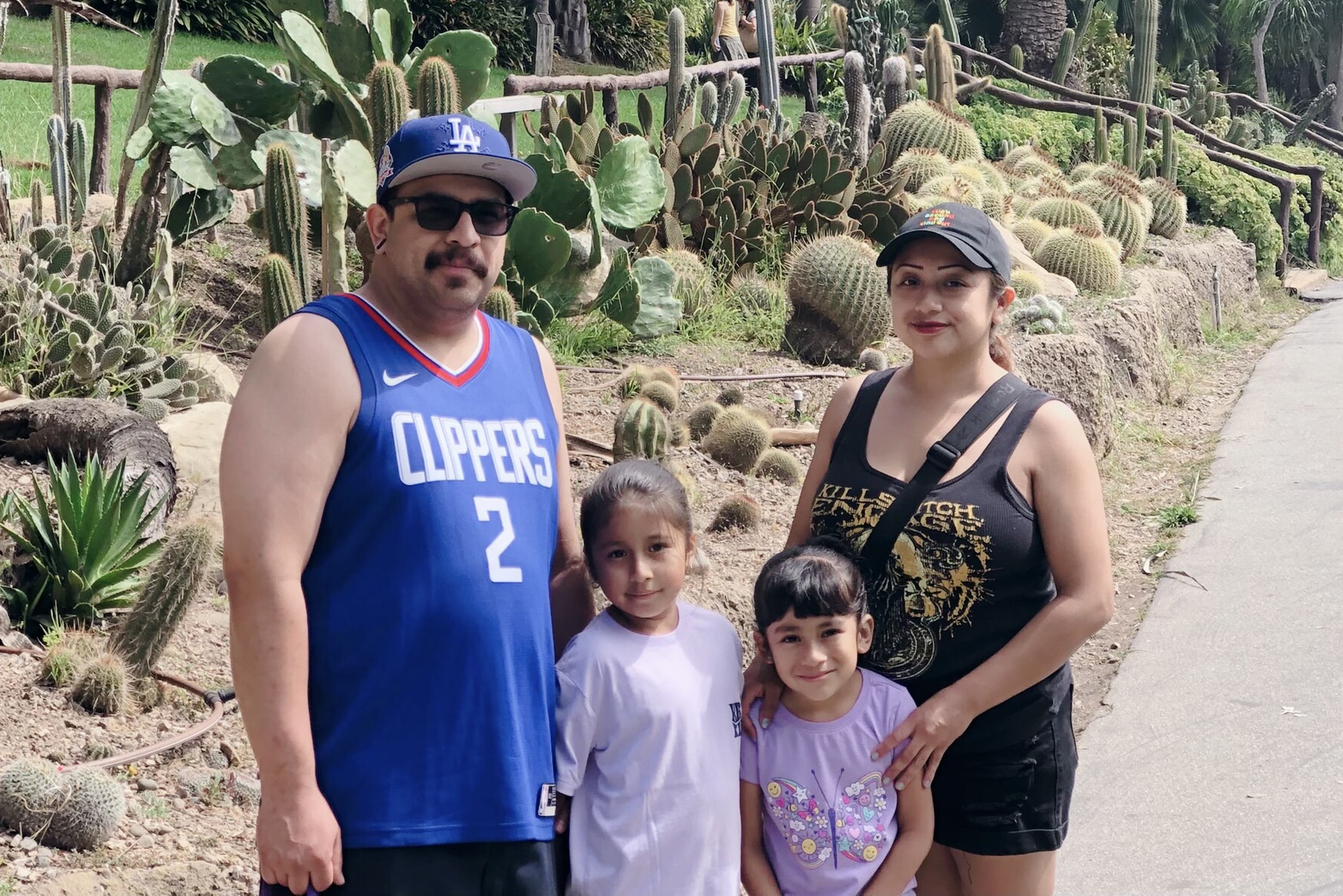 A Family Of Four Poses On A Path Next To A Cactus Garden. The Adults Wear Hats And Casual Summer Clothes, While The Two Young Girls Stand In Front, Smiling. It Is A Sunny Day.