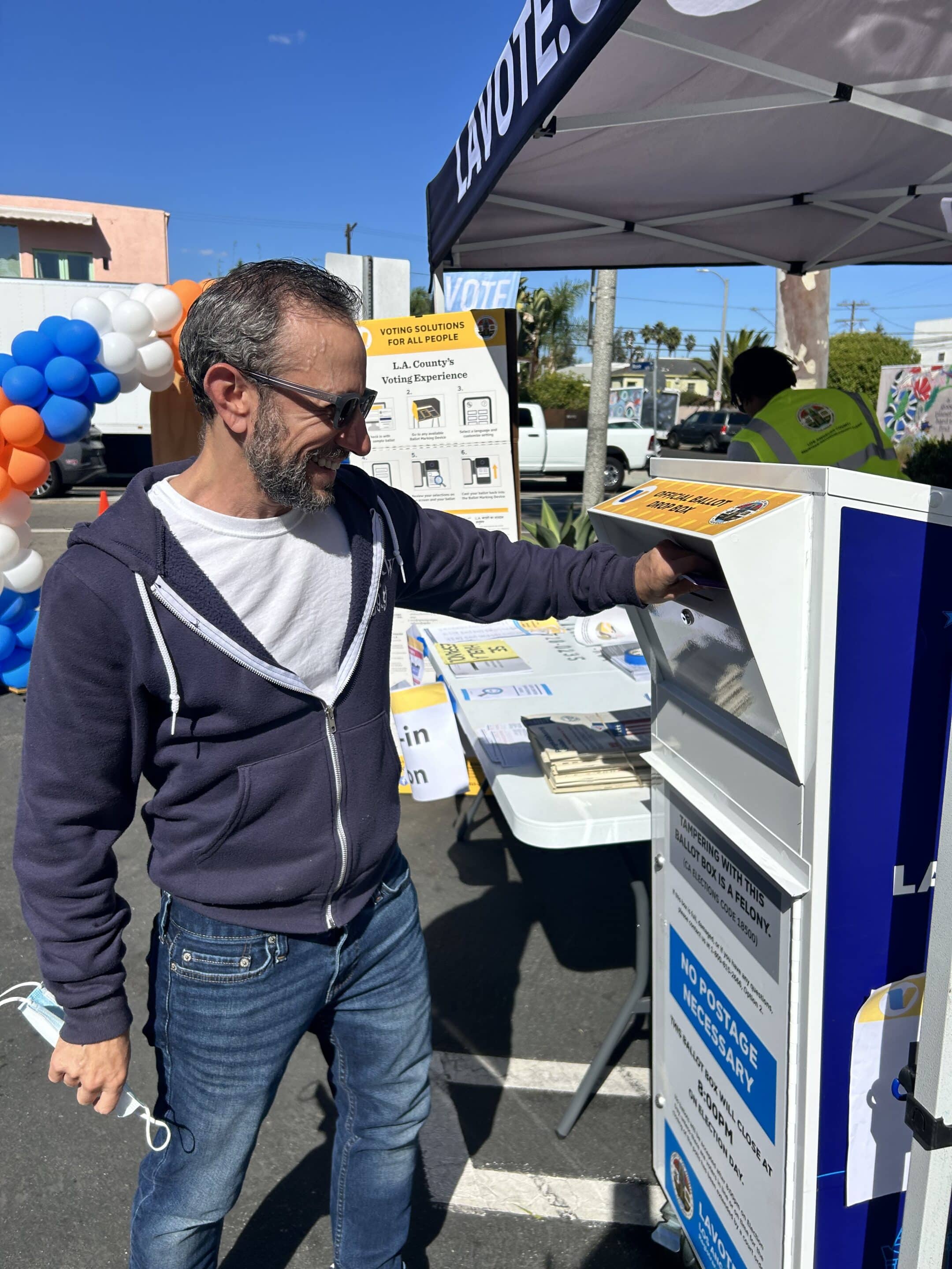 A Man Wearing Sunglasses And A Hoodie Places A Ballot In An Official Ballot Drop Box At An Outdoor Voting Event Under A Tent, With Informational Signs And Balloons In The Background.