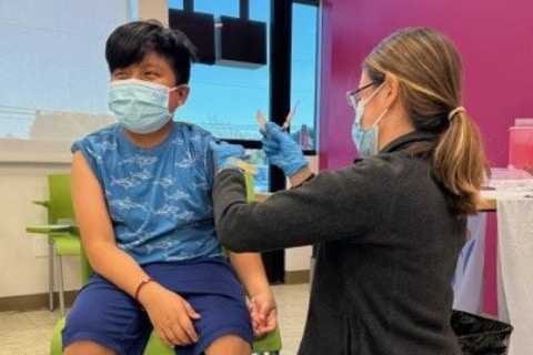 A Child Wearing A Mask Sits As A Healthcare Worker, Also Masked And Gloved, Prepares To Give Him A Vaccine Shot In The Arm In A Clinic Setting. A Child Wearing A Mask Sits As A Healthcare Worker, Also Masked And Gloved, Prepares To Give Him A Vaccine Shot In The Arm In A Clinic Setting.