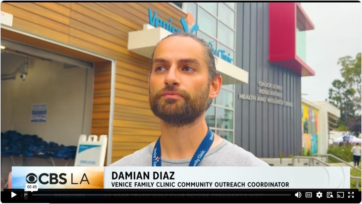 A Man With A Beard And Tied-back Hair Stands Outside A Clinic Building; Text Reads CBS LA, Damian Diaz, Venice Family Clinic Community Outreach Coordinator.. A Man With A Beard And Tied-back Hair Stands Outside A Clinic Building; Text Reads CBS LA, Damian Diaz, Venice Family Clinic Community Outreach Coordinator..