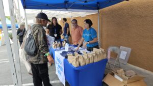 Four People Interact At An Outdoor Booth Under A Blue Tent. The Table Displays A Native Health Sign And Many Brown Paper Bags. One Woman Hands Out Items While Others Stand And Watch.