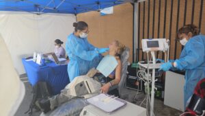A Patient Receives Dental Care From A Masked Provider Under A Blue Tent, While Another Provider Uses Medical Equipment Nearby And Two People Work On Laptops At A Table In The Background.