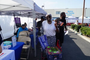 A Woman Smiles And Flashes A Peace Sign While Standing Outside Near Tents And Tables At A Community Event. Other People Are Nearby, And A Venice Family Clinic Sign Is Visible In The Background.