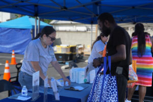 A Woman At An Outdoor Event Table Hands Paperwork To A Man Holding A Blue Bag. Other People Are Nearby Under Blue Tents. Safety Cones And Supplies Are Visible In The Background.