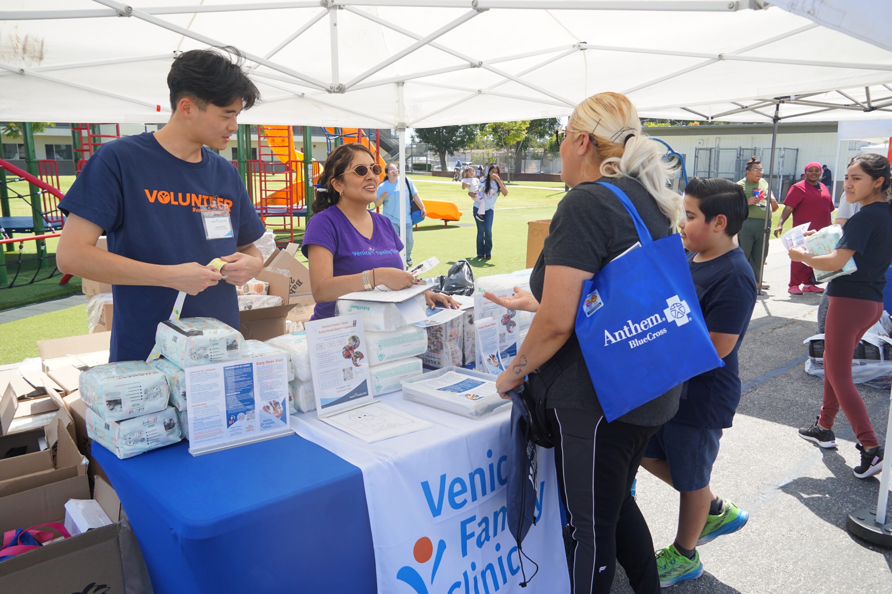 Volunteers At An Outdoor Booth Hand Out Diapers And Information To A Woman And Child. The Booth Displays Venice Family Clinic Materials, And People Are Engaged In Conversation Under A White Canopy. Volunteers At An Outdoor Booth Hand Out Diapers And Information To A Woman And Child. The Booth Displays Venice Family Clinic Materials, And People Are Engaged In Conversation Under A White Canopy.