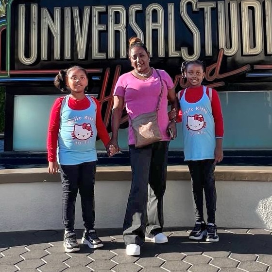 A Woman Stands Between Two Girls In Matching Hello Kitty Shirts In Front Of A Universal Studios Hollywood Sign, All Smiling At The Camera.