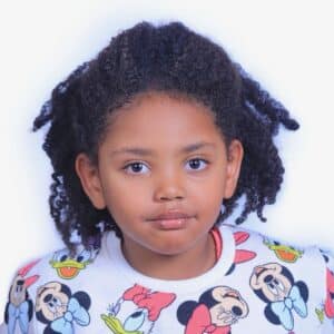 A Young Child With Curly Hair Wearing A White Sweatshirt Featuring Colorful Cartoon Characters, Including Mickey Mouse And Donald Duck, Looks Directly At The Camera Against A Plain White Background.