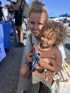 A Smiling Woman Kneels Next To A Young Child Holding Colorful Toys At An Outdoor Event, With People, Tables, And Tents Visible In The Background Under A Clear Blue Sky.