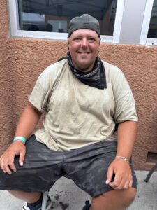 A Man In A Tan Shirt, Camo Shorts, And A Backward Cap Sits On A Stool Outside Against A Textured Wall, Smiling At The Camera. He Wears A Black Bandana Around His Neck And A Wristband On His Right Wrist.
