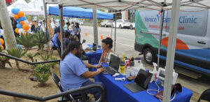 Two People Work At Laptops Under A Tent Beside A Venice Family Clinic Van. Medical Equipment And Supplies Are On The Tables. Balloons And People Are Seen In The Background, Suggesting A Community Health Event.