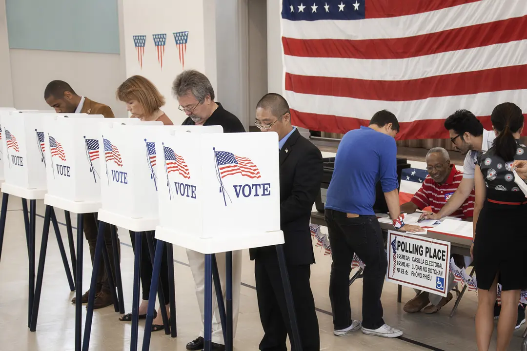 People Cast Ballots At Private Voting Booths And A Registration Table In Front Of A Large American Flag.