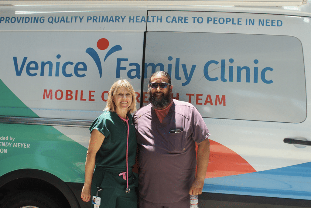 A Woman And A Man In Medical Scrubs Stand In Front Of A Venice Family Clinic Mobile Van.